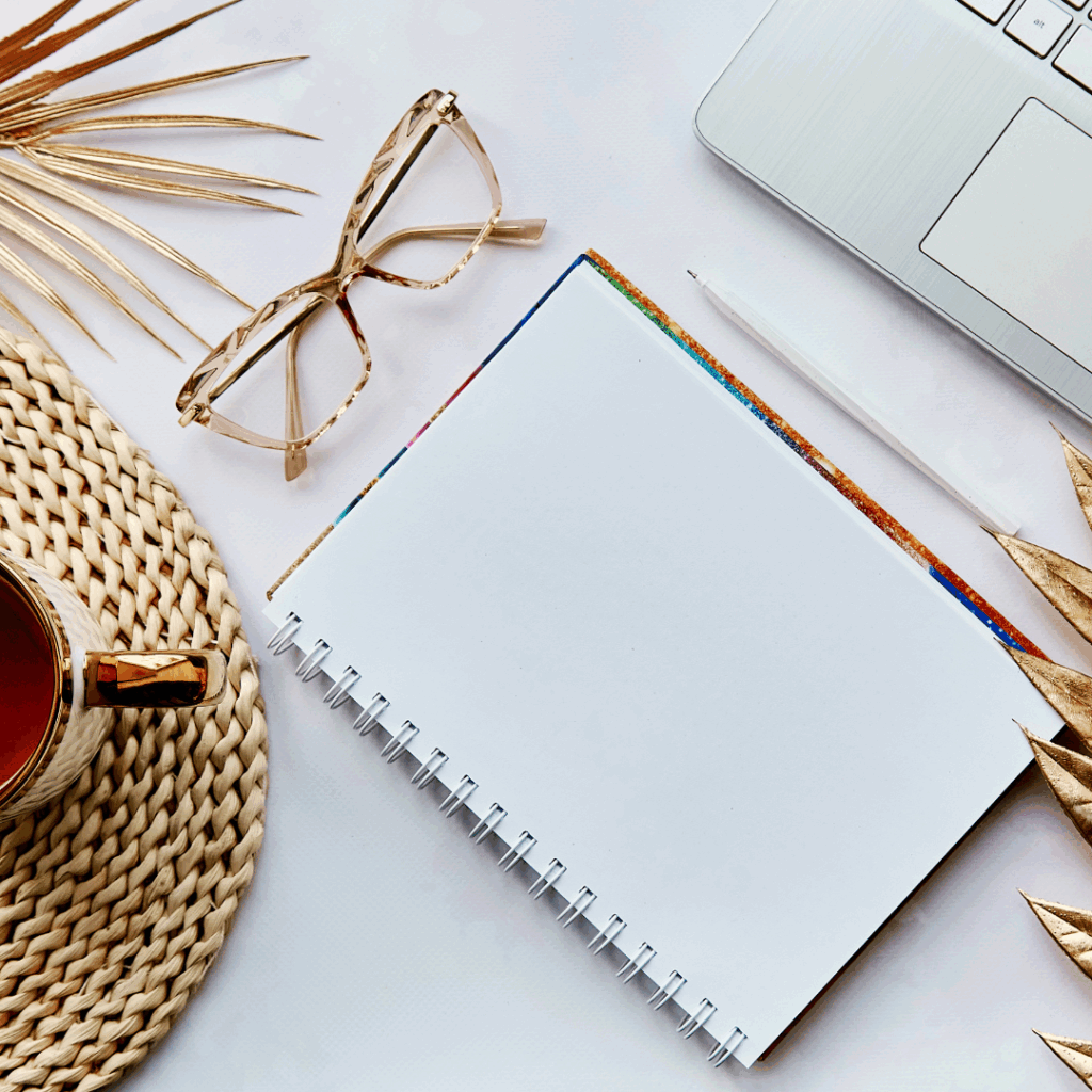 Flat lay of notebook, glasses, and laptop on a desk with gold accents and tea