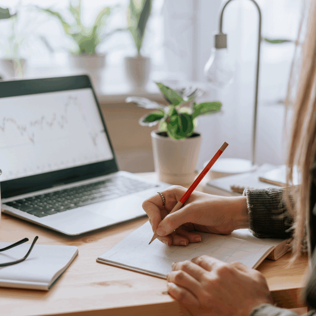 Person writing in a planner next to a laptop displaying a graph, surrounded by plants and natural light