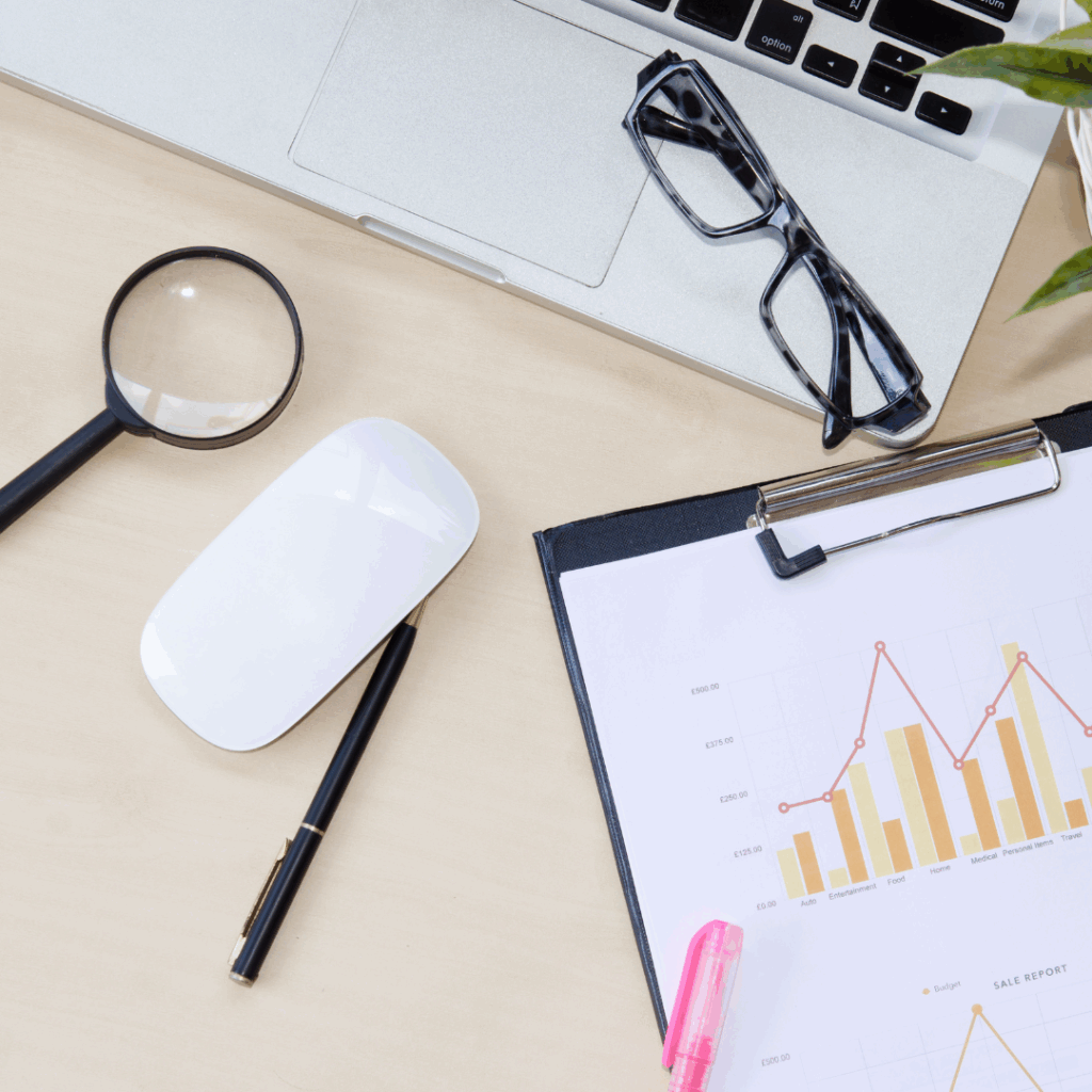 Desk with laptop, magnifying glass, chart clipboard, and glasses representing real estate pricing analysis