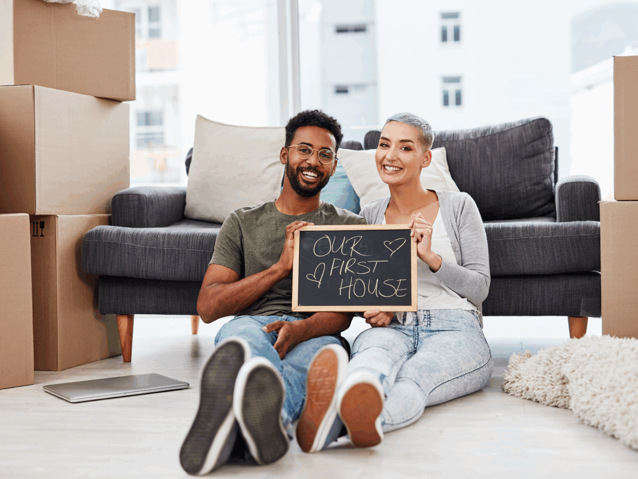 Happy couple sitting on floor with moving boxes, holding chalkboard sign that says "Our First House"